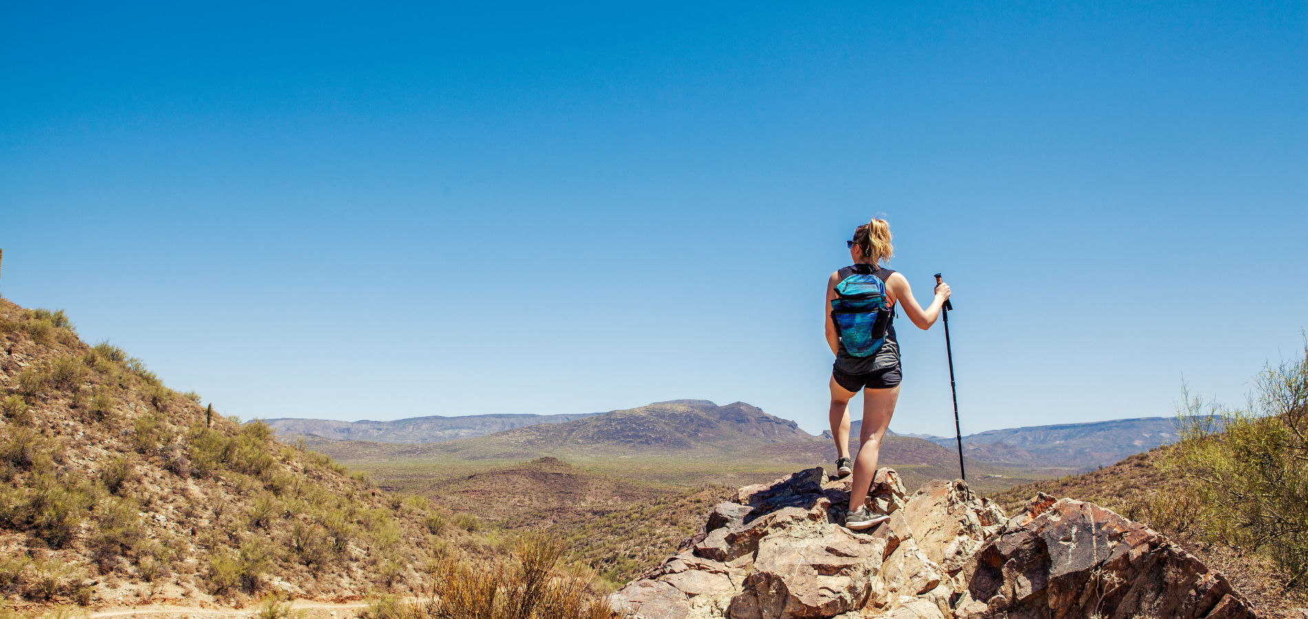 Hiker with backpack and trekking pole standing atop rocky terrain overlooking vast desert landscape under a blue sky.